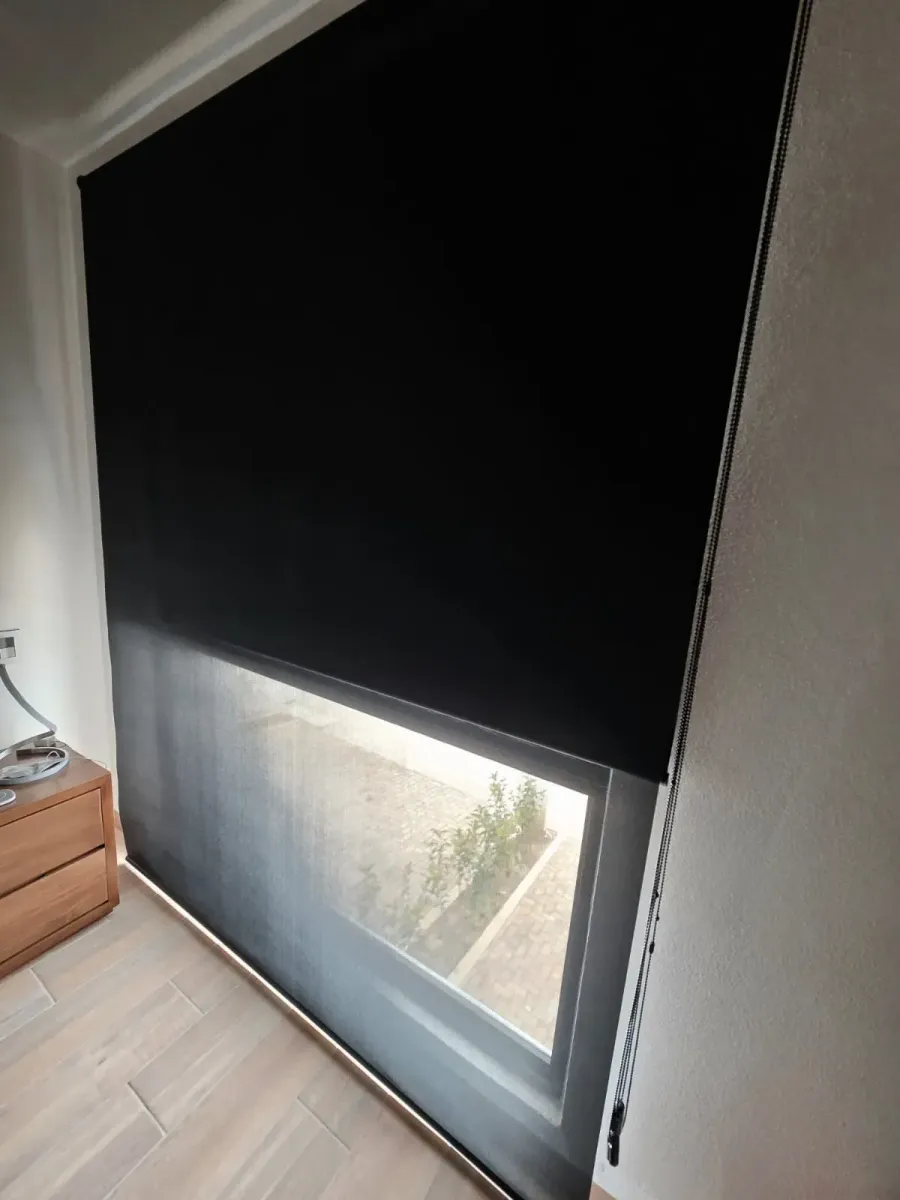 Persiana enrollable blackout negra a medio bajar en una recámara con piso de madera